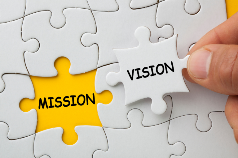 A close-up of a puzzle piece with the words "VISION" and "MISSION" printed on it in black letters. The puzzle piece has a slightly textured, yellow background. community builders